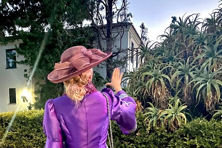 Person in einem lila Kleid und Hut steht im Freien in der Nähe von großen Aloe-Pflanzen.